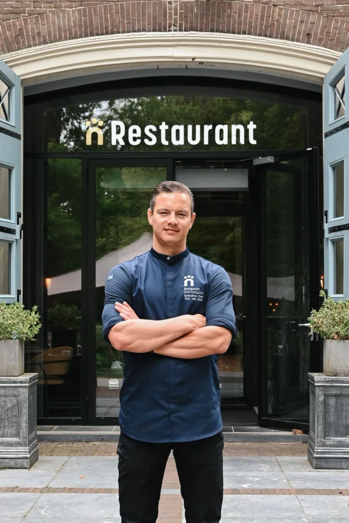 Roy van Geel in koksbuis met gekruiste armen voor de ingang van het restaurant