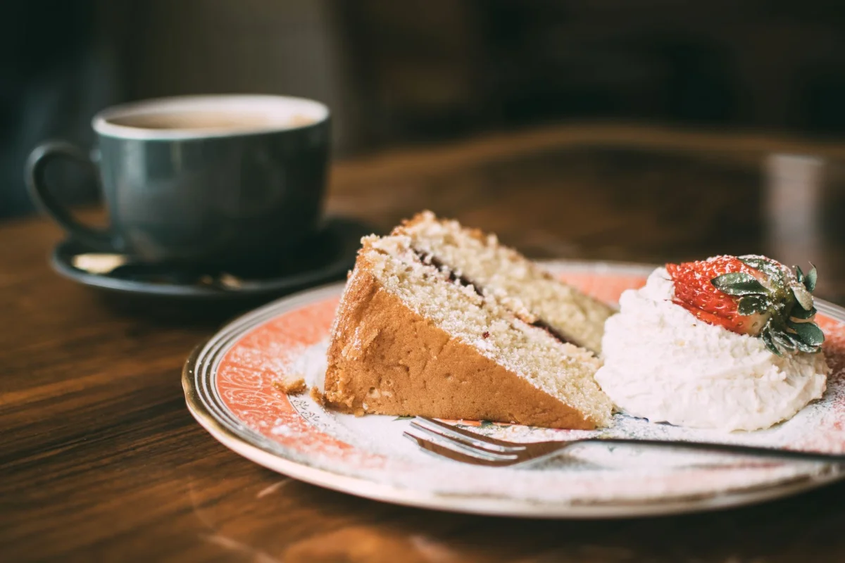 A slice of cake with cream and strawberry beside a cup of coffee